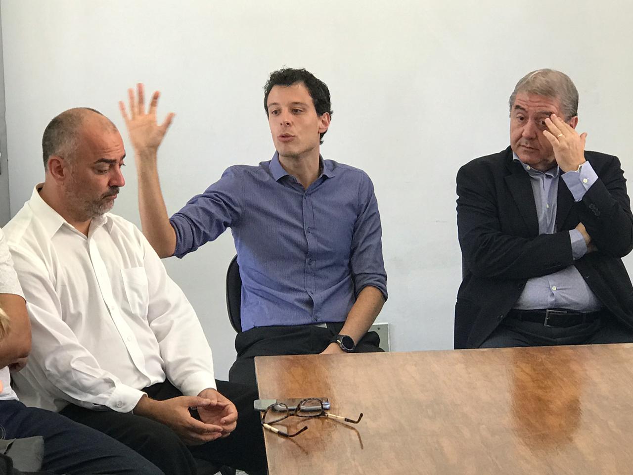 Três homens e um deles falando com uma das mãos levantadas em sinal de expressão da sua fala 